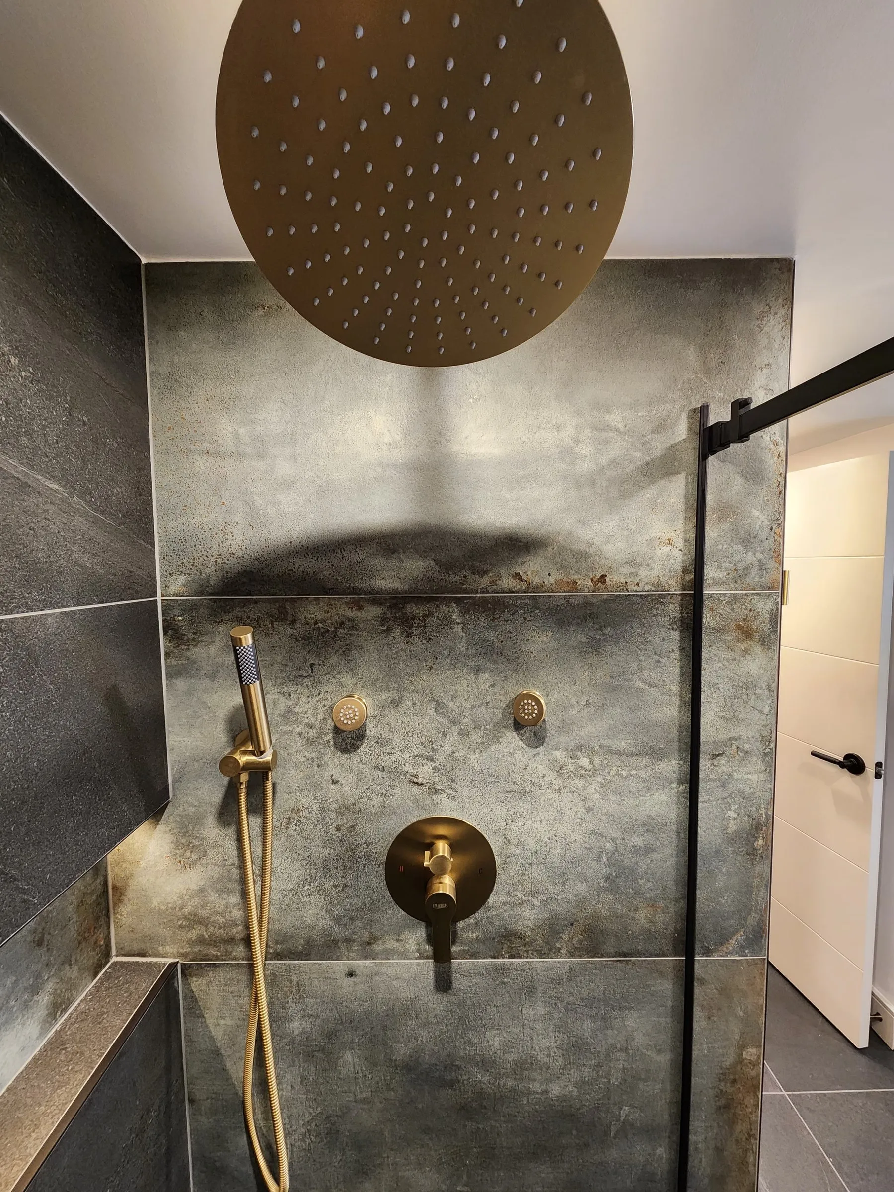A shower with large dark stone-look tiles, a round brass rain shower head and brass controls