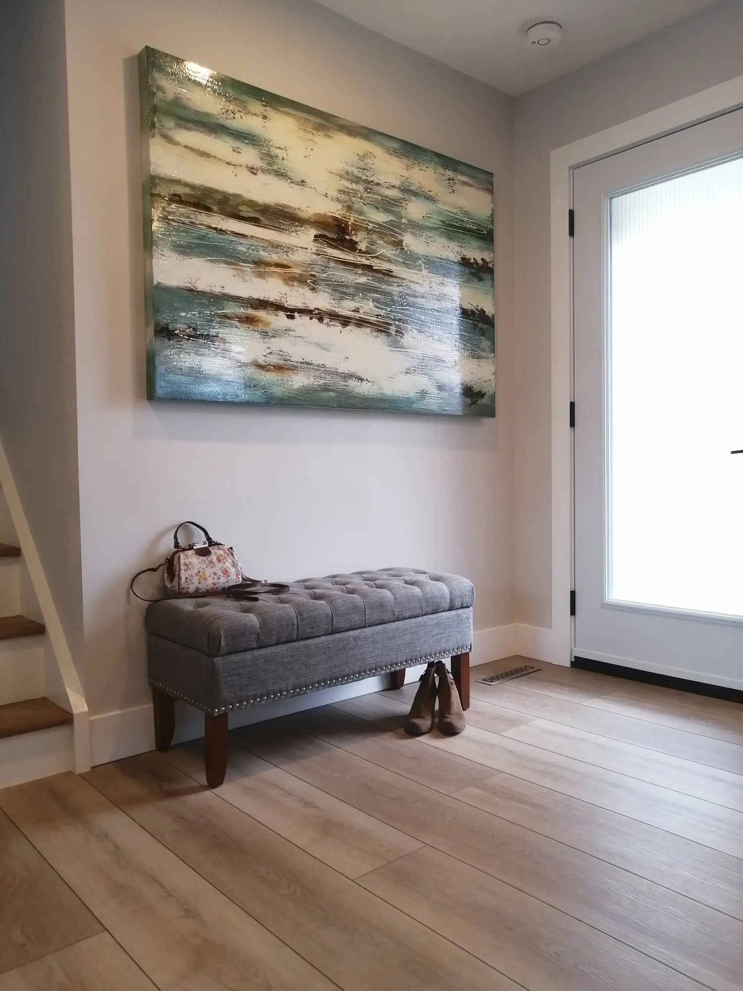 A finished front entryway with a grey tufted bench, large abstract painting, light plank floor and white door.
