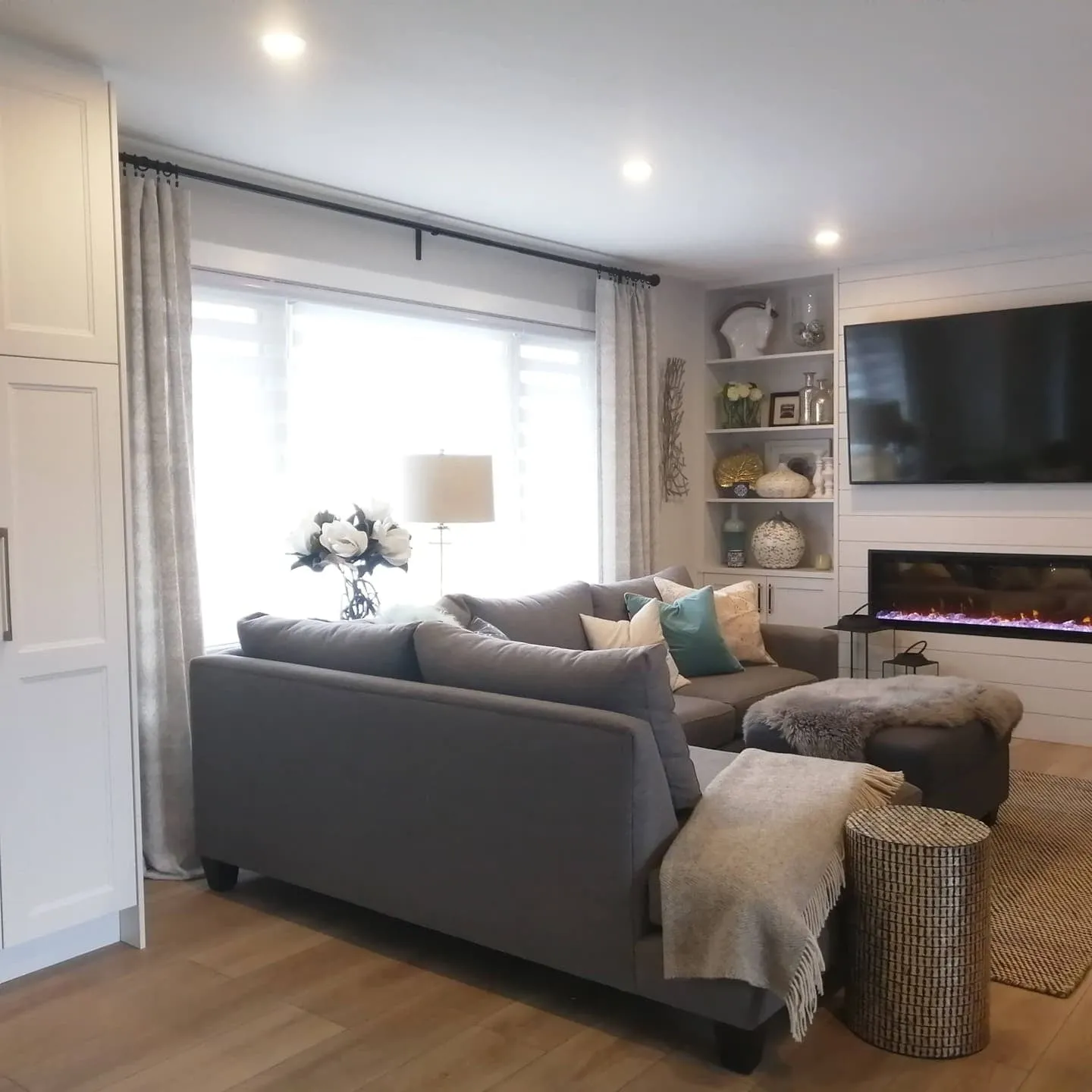 A bright living room with a grey sectional, a large curtained window and a white shiplap fireplace wall.