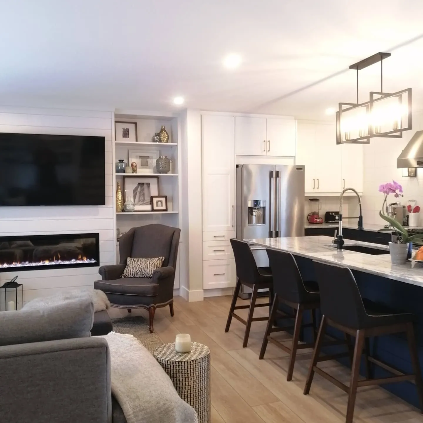 An open living room and kitchen with white cabinets, stainless fridge and a dark island with three stools.