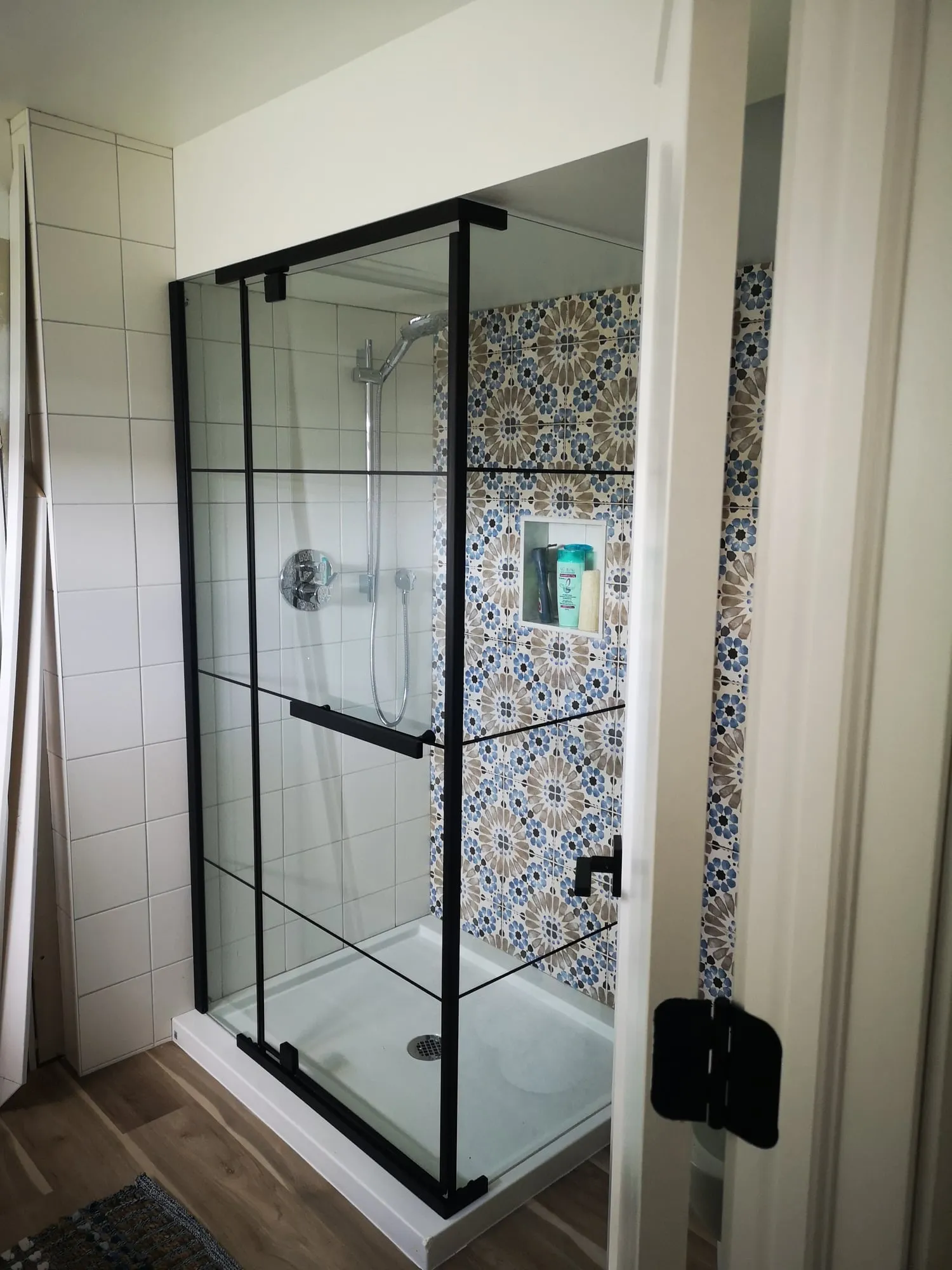 A walk-in shower behind black-framed glass, with white tile and a blue-and-tan patterned wall with a niche.