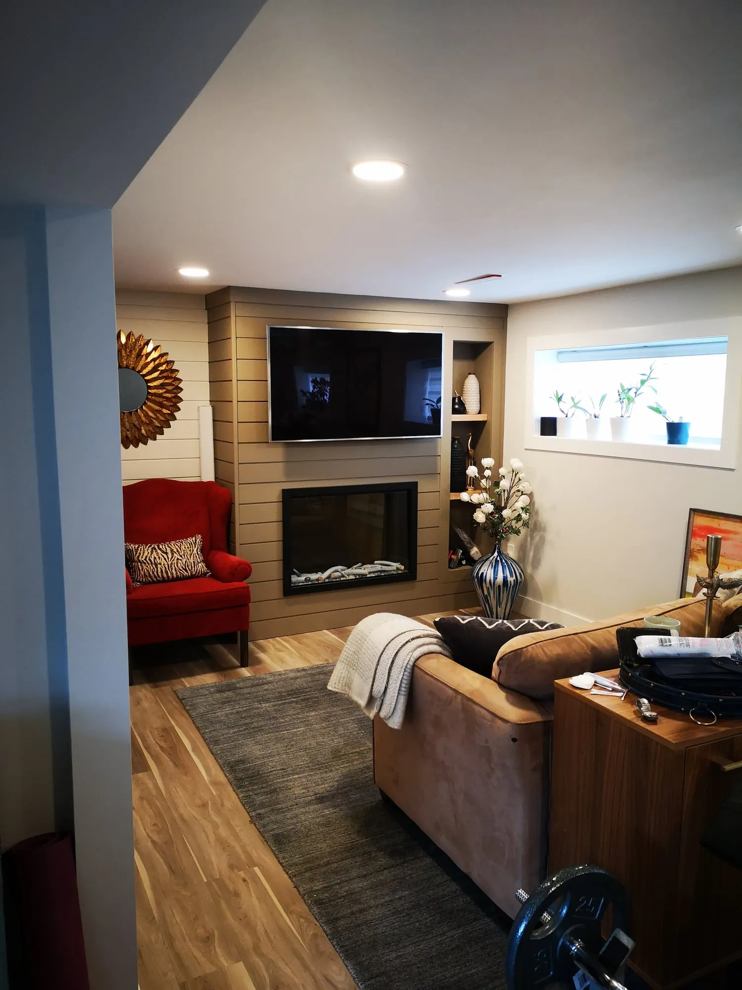 A cosy family room with a taupe shiplap fireplace wall, mounted TV, red wingback chair and tan sofa.