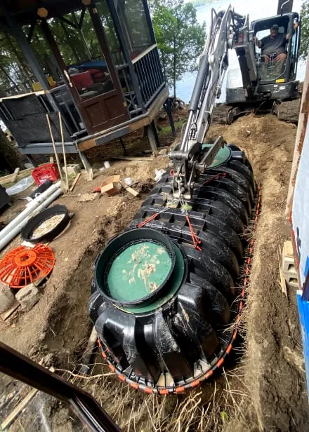 A new black septic tank set into an open trench beside the cottage, with a small excavator behind.