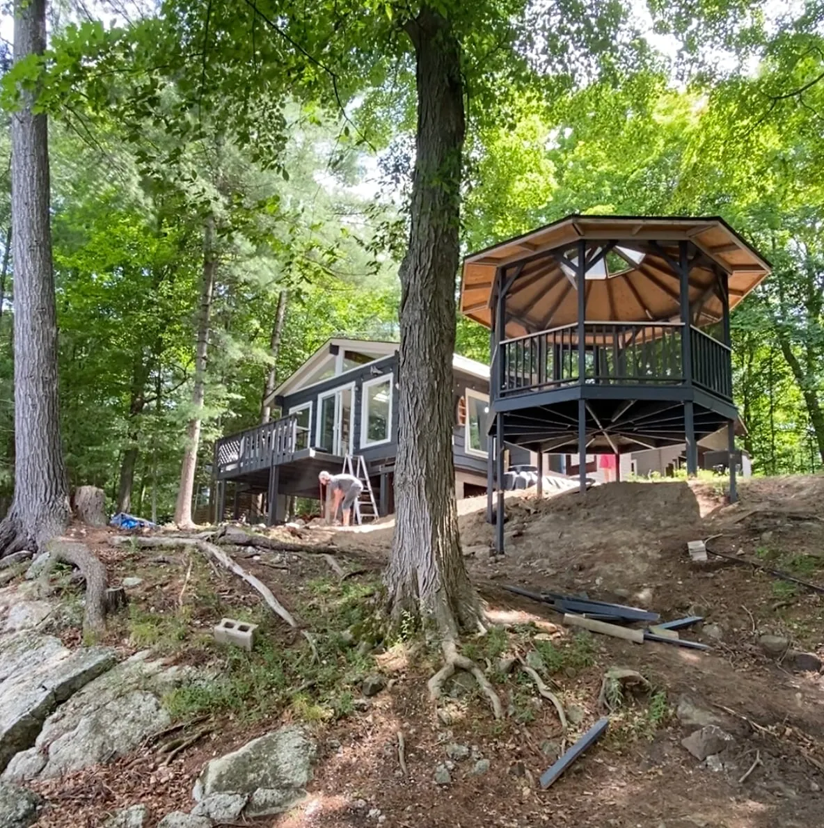 The octagonal black gazebo on tall posts beside the cottage before removal, set on a wooded slope.