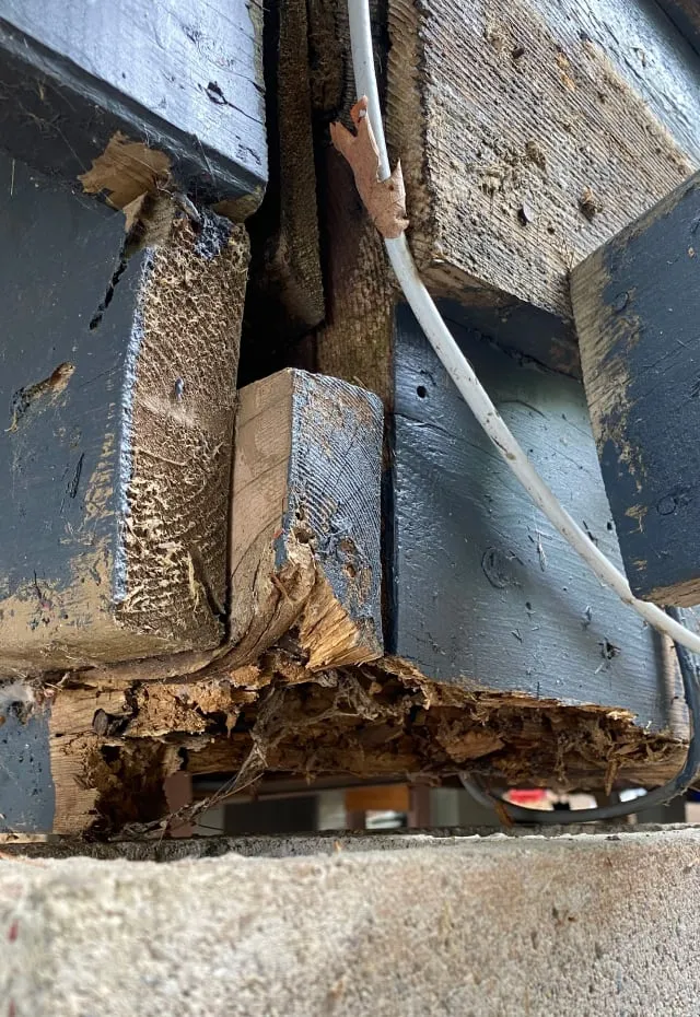 A close-up of the old deck's support, with dark painted beams and crumbling, rotted wood at the base.