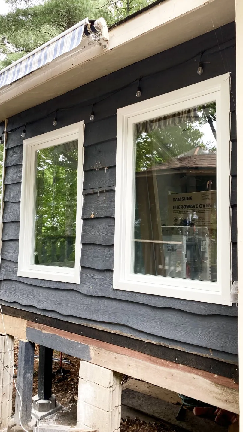 Two new white-framed windows in the cottage's dark blue-grey siding, with string lights along the eaves.