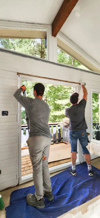 Two workers reaching up to the top of a tall opening in a white panelled wall, under gable windows.