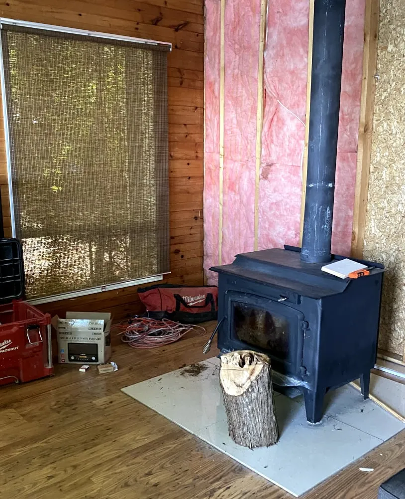 The old black wood stove on a tile hearth, with pink insulation exposed in the stripped wall behind.