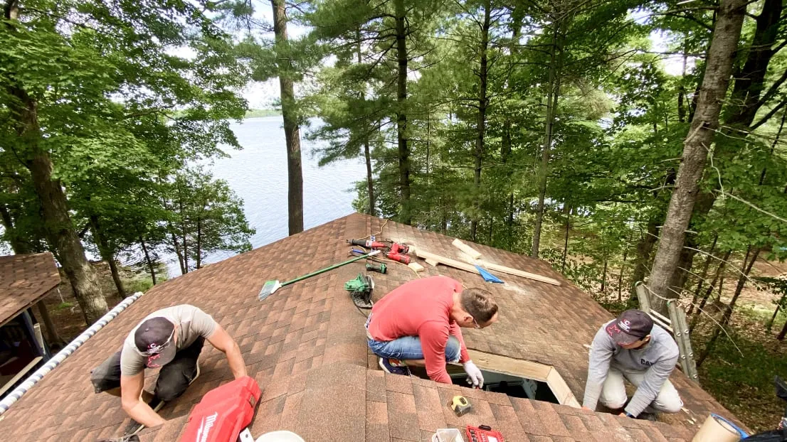 Three workers on a brown shingled roof around a skylight opening, with the lake beyond the trees.