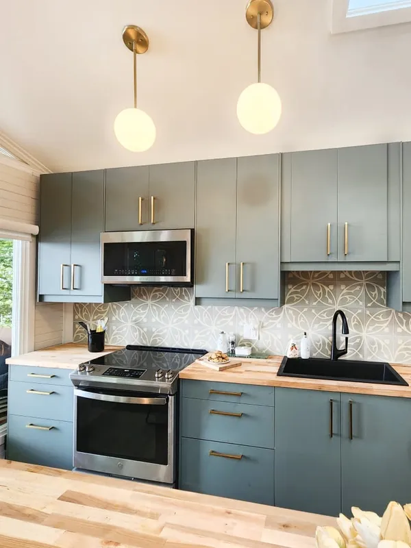 The finished kitchen with sage green cabinets, gold handles, a patterned tile backsplash, black sink and globe pendants.
