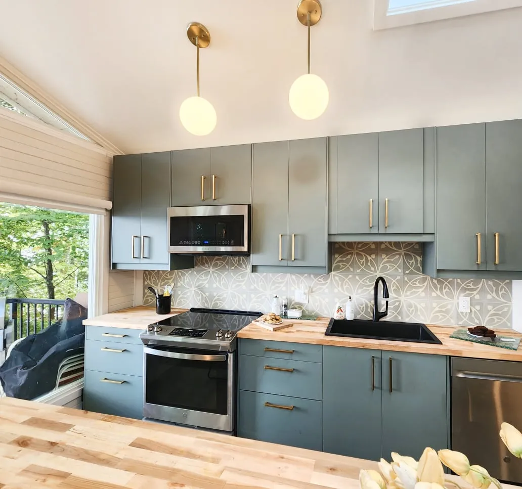 The finished kitchen with sage green cabinets, gold handles, a patterned tile backsplash, black sink and globe pendants.