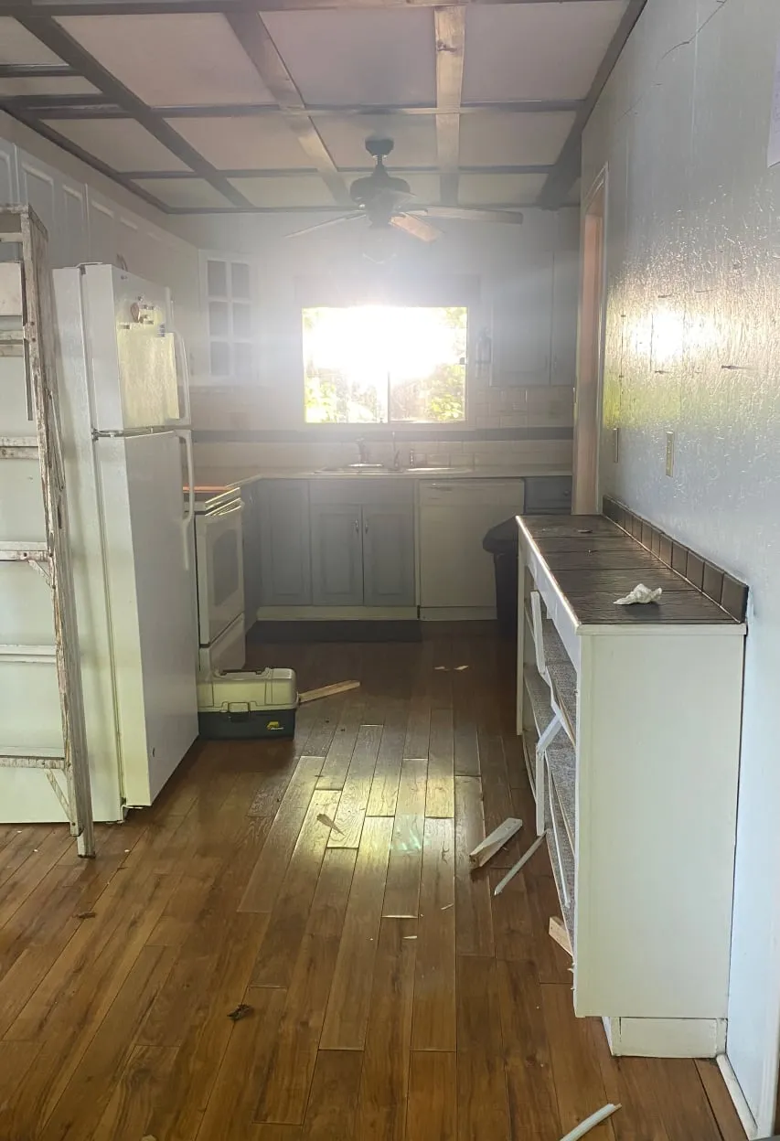 The old narrow kitchen with a white fridge and stove, grey cabinets and a window above the sink.