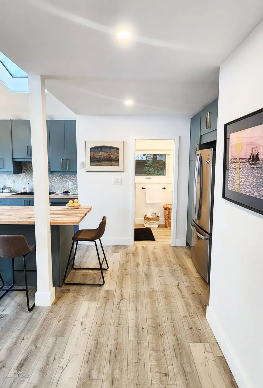 A walkway with light plank floors between the kitchen island and a stainless fridge, toward an open doorway.