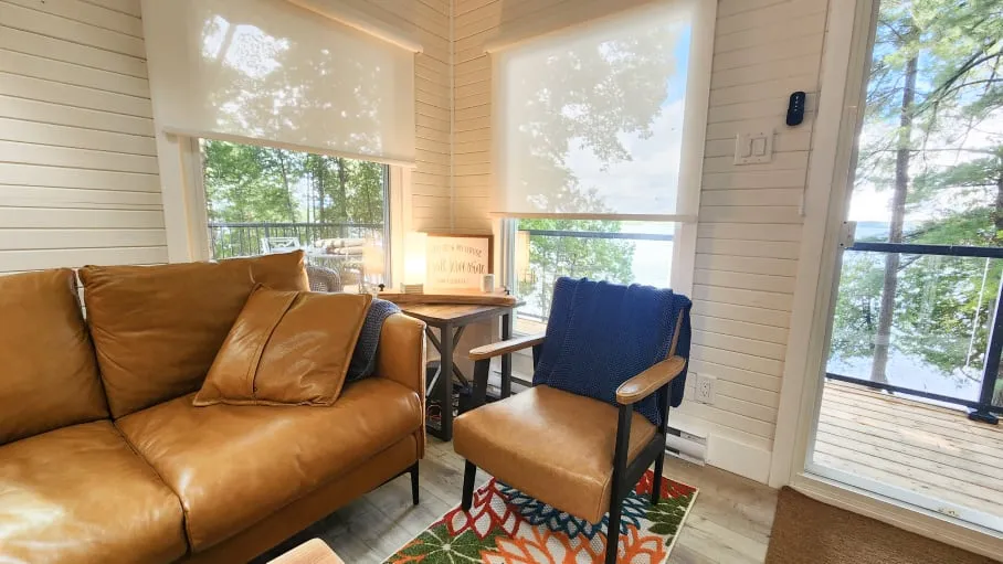 A corner of white panelled walls and shaded windows, with a tan sofa, armchair and colourful rug.
