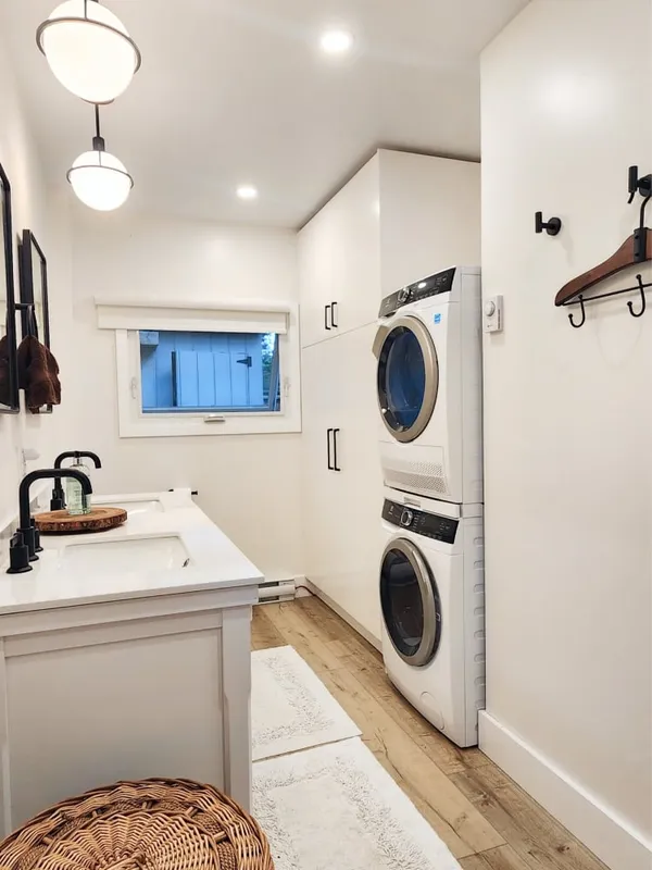The finished bathroom with stacked white laundry machines, tall white cabinets, two globe pendants and wood-look floors.