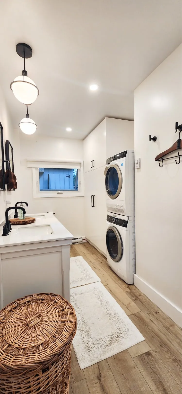 The finished bathroom with stacked white laundry machines, tall white cabinets, two globe pendants and wood-look floors.