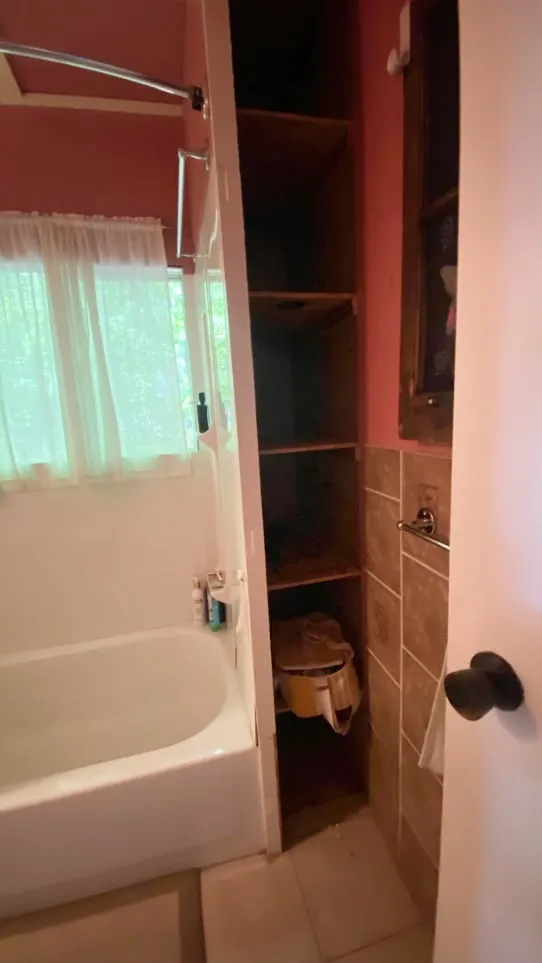 The old bathroom's narrow open shelves squeezed between the tub and a pink wall with brown tile.