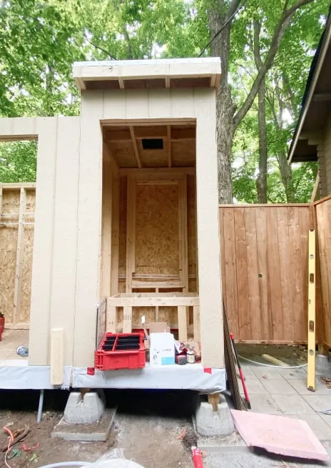 A small beige-sided outbuilding under construction, its open front showing bare wood sheathing and framing inside.