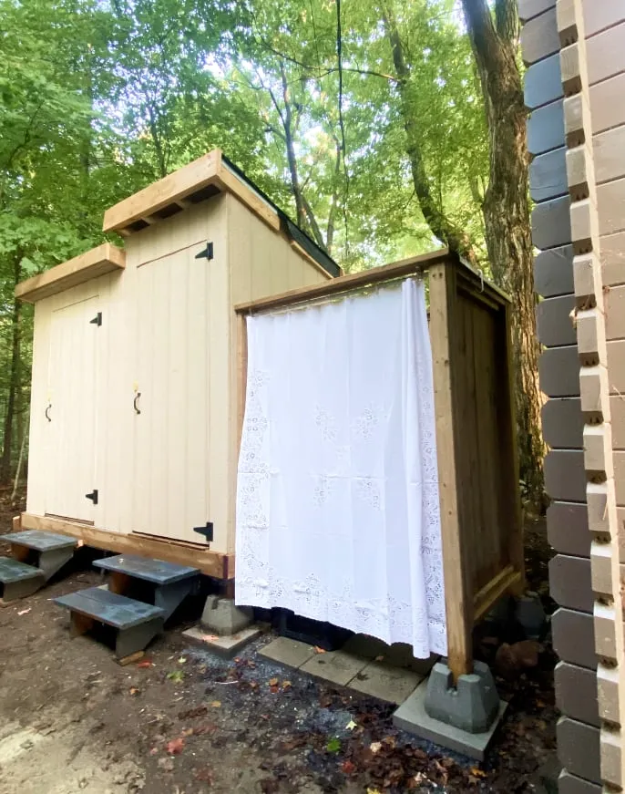 A beige two-door outbuilding beside a wooden outdoor shower with a white lace curtain, among tall trees.