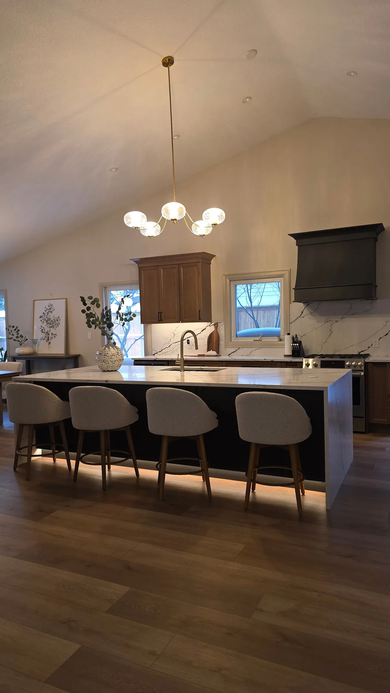 The kitchen under a vaulted ceiling: a long island with four upholstered stools, a globe chandelier above and a black range hood beyond.