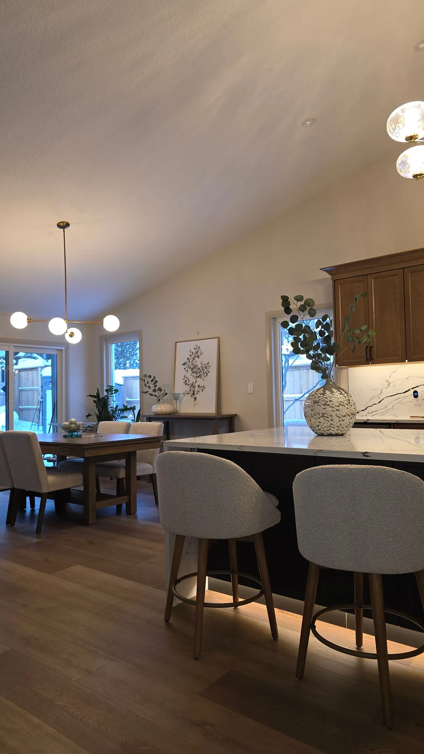 Stools along the island with soft light glowing beneath it, and the dining table and windows beyond.