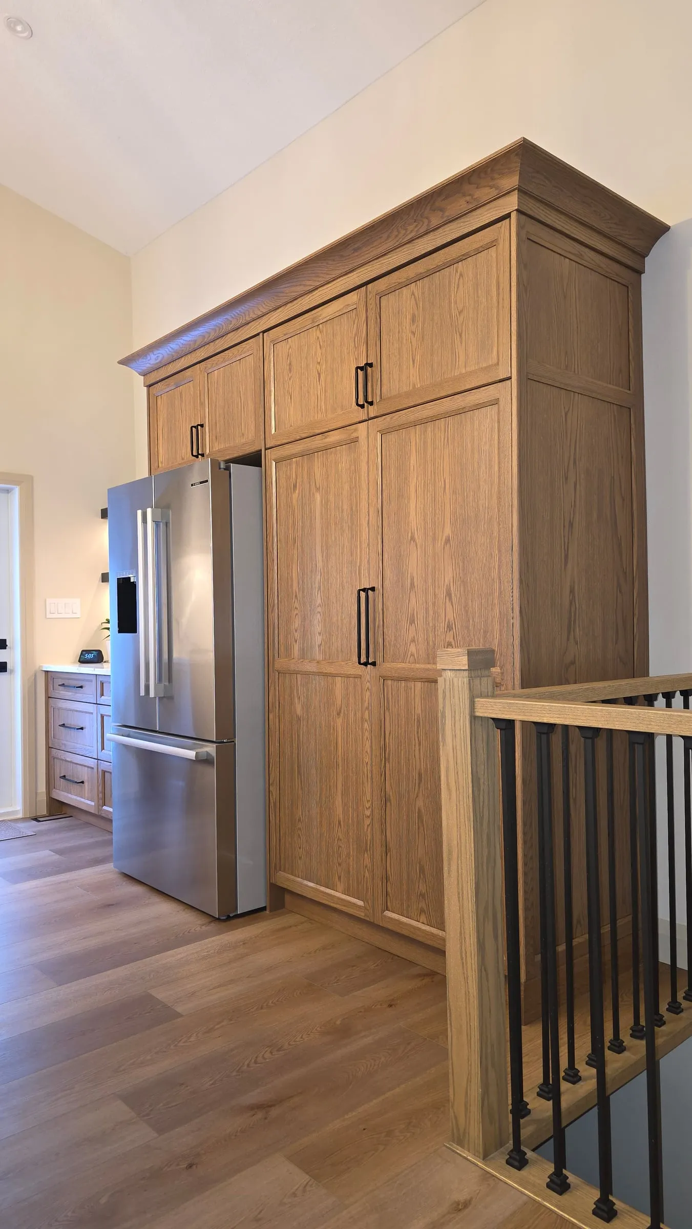 Tall wood cabinets with crown moulding above and beside a stainless fridge, next to the stair railing.