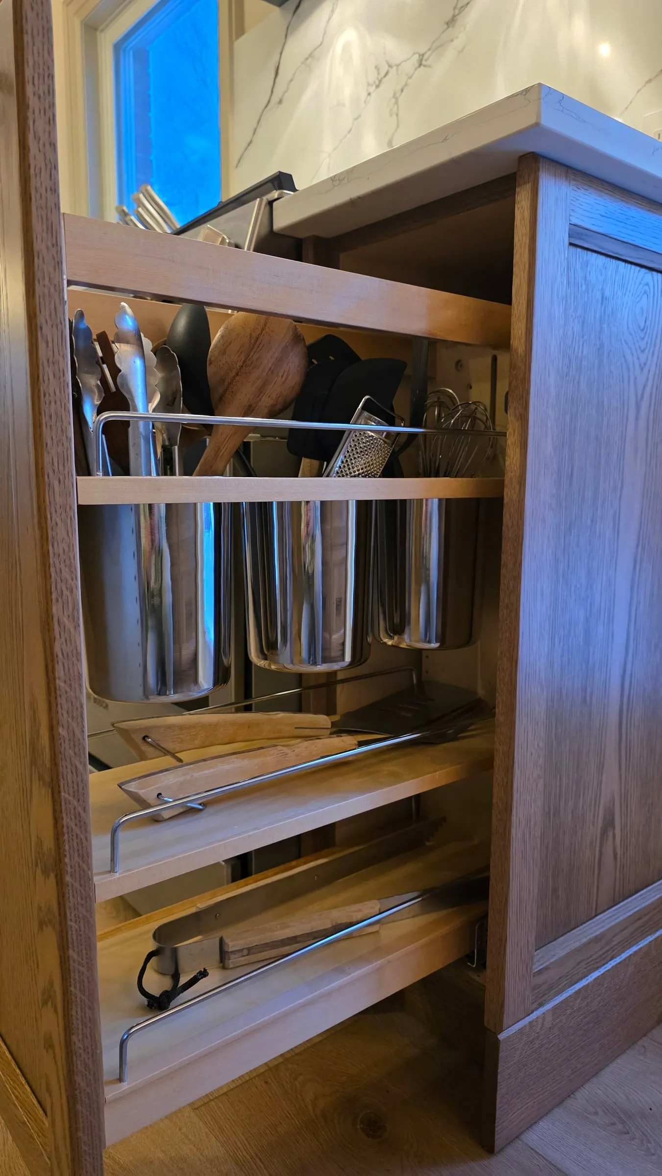 A pull-out organizer holding utensils in steel cups, with cutting boards, a spatula and tongs on the shelves below.
