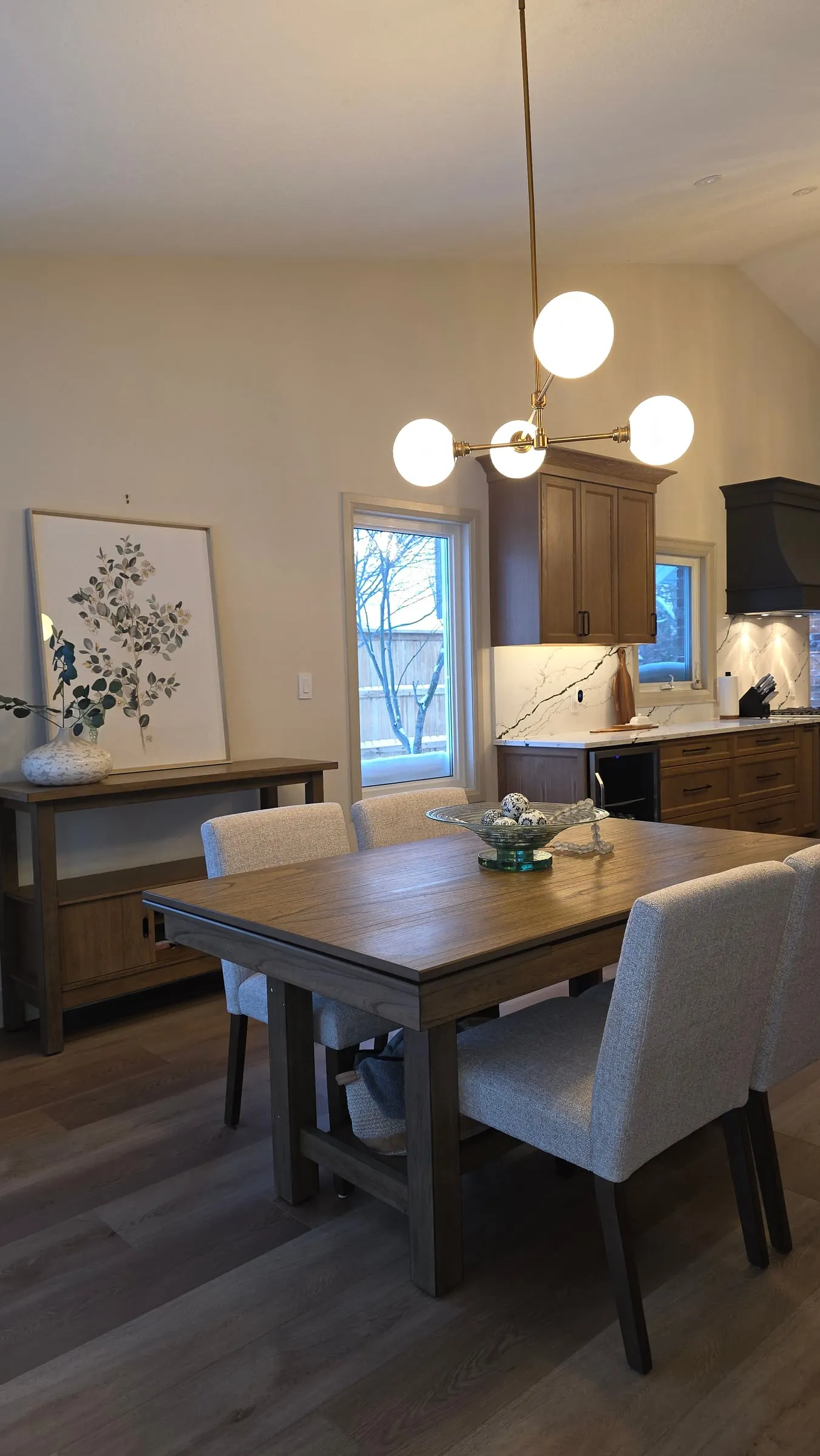 A wood dining table with upholstered chairs under a brass globe light, with the kitchen in the background.