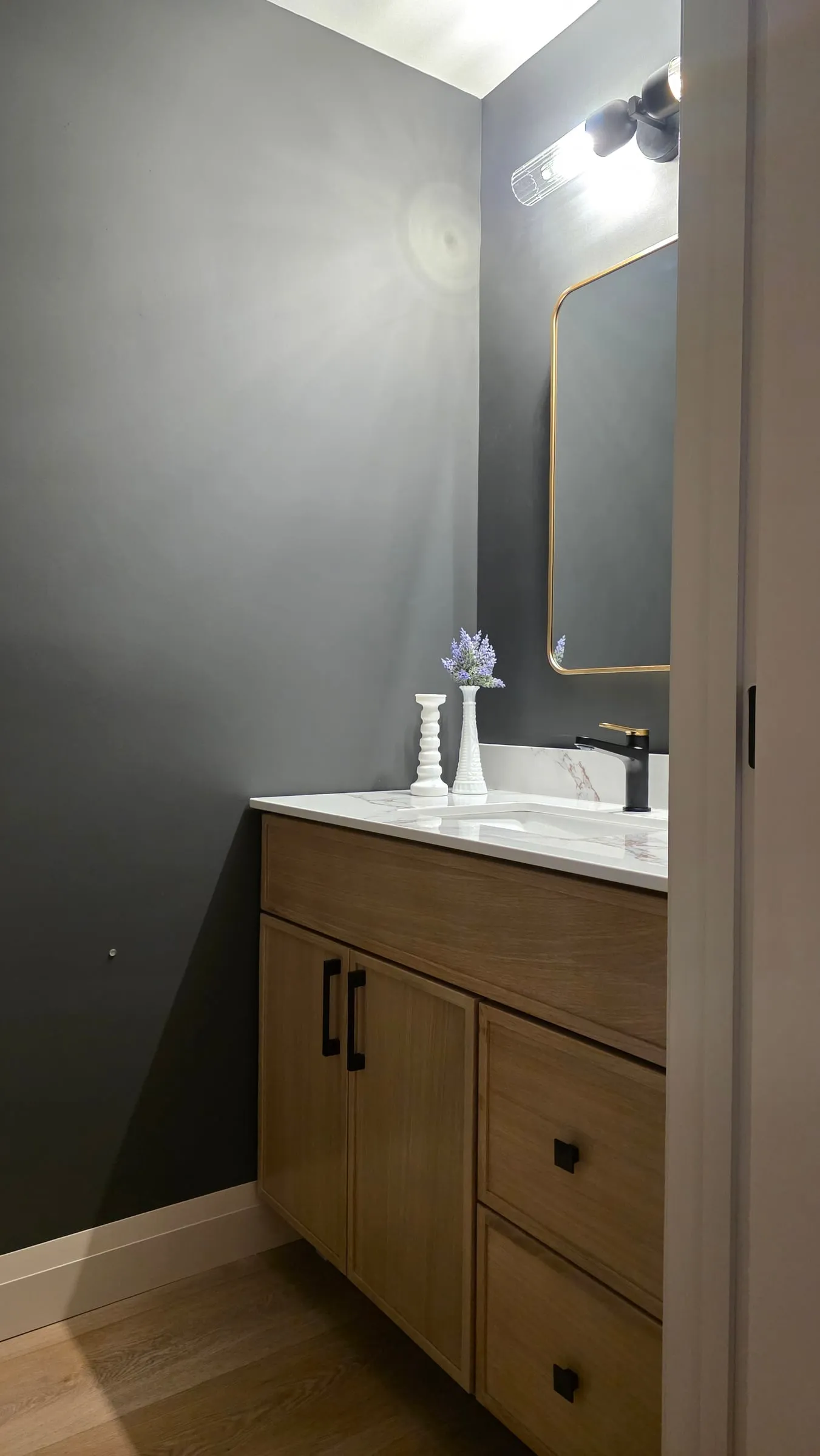 A bathroom with charcoal walls, a wood vanity with a white top and a black faucet, and a brass-framed mirror.